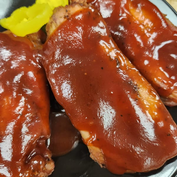 Three BBQ ribs, generously coated in a rich, glossy sauce, with yellow pickled peppers on the side.