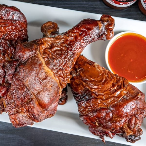 Three glistening, dark brown BBQ turkey legs on a white platter with a side of red sauce.