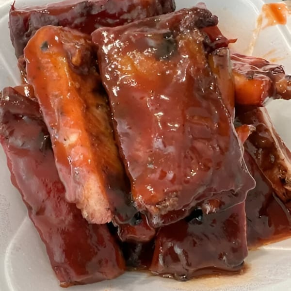 Close-up of several glistening, saucy BBQ ribs from Fat Man's BBQ in a white container.